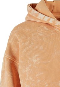 Felpa arancione con una consistenza morbida, caratterizzata da un sottile motivo tie-dye e un cappuccio ampio. Il tessuto è leggero ma caldo.