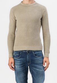 Homme portant un pull en tricot texturé beige et un jean bleu délavé, debout devant un fond blanc uni.