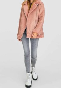 Lichtroze nepvachtjas met sjaalkraag, ritssluiting en zijzakken, gecombineerd met grijze jeans en witte veterlaarzen.