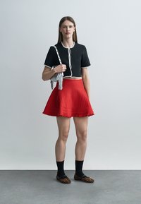 Młoda kobieta ubrana w czarną bluzkę z krótkim rękawem, czerwoną plisowaną spódnicę, czarne skarpetki i baleriny w panterkę, trzymająca srebrną torebkę na ramieniu.