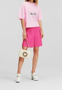 Osoba oblečená v volném růžovém tričku Karl Lagerfeld, jasně růžových šortkách, bílých platformových botách a držící béžovou kabelku s kruhovým zlatým logem.