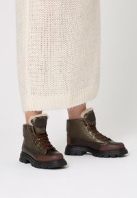 Olivově zelené kotníkové boty s hnědými koženými detaily, umělou kožešinou, hnědými tkaničkami a černou robustní podrážkou, umístěné na světlém pozadí.