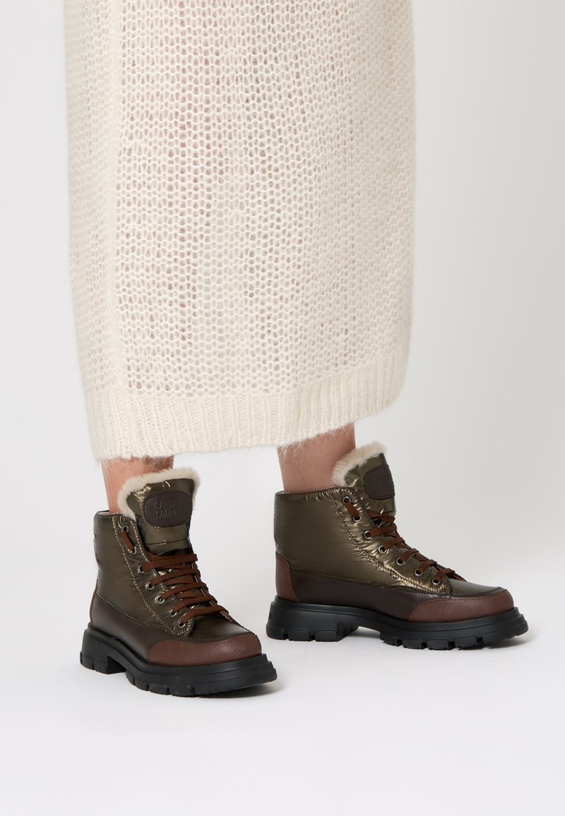 Olivově zelené kotníkové boty s hnědými koženými detaily, umělou kožešinou, hnědými tkaničkami a černou robustní podrážkou, umístěné na světlém pozadí.
