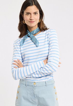 Femme aux cheveux foncés portant des boucles d'oreilles anneaux dorés, un foulard bleu à motifs autour du cou, une chemise à manches longues à rayures bleu clair et blanc, et un pantalon rayé bleu.