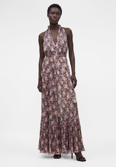 Γυναίκα με αμάνικο, μακρύ φόρεμα χρωματιστό με λεπτή ζώνη, στέκεται με το ένα χέρι χαλαρό, σε λευκό φόντο.