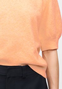 Κοντινή λήψη προσώπου που φοράει μάλλον κοντομάνικο ροδακινί πουλόβερ στερεωμένο μέσα σε σκούρο μπλε παντελόνι με ορατό θηλάκι ζώνης.