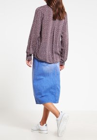 Brunmönstrad långärmad blus med rynkade ärmar, tillsammans med en blå denim-midi kjol och vita sneakers. Synlig bak sömdetalj.