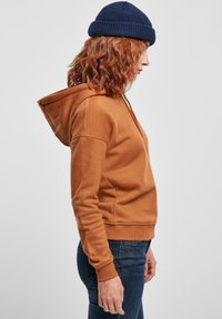 Smeđi pamučni hoodie s kapuljačom, džepom tipa kangaroo, rebrastim manžetama i donjim rubom. Uparen s tamnim trapericama i mornarskom pletivom kapom.