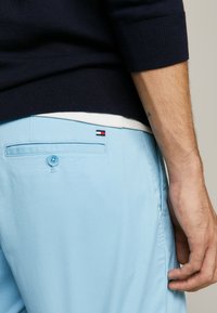 Pantaloni corti blu chiaro con chiusura a bottone, dotati di una tasca posteriore e di una piccola etichetta logo rossa, bianca e nera. Maglione blu navy visibile.