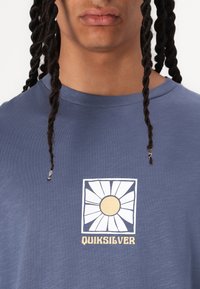 Tricou bleu cu maneci scurte, confecționat dintr-un material moale, care prezintă un grafic floral alb cu un centru galben și textul "Quiksilver" în partea de jos.