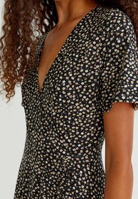 Robe noire avec un motif floral comportant des fleurs blanches et jaunes. Manches courtes et décolleté en V profond. Tissu léger avec une texture douce.