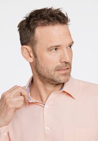Camisa abotonada en color melocotón claro con un acabado texturizado y un forro de cuello con un patrón floral. Presenta un bolsillo en el pecho y un cuello estándar.