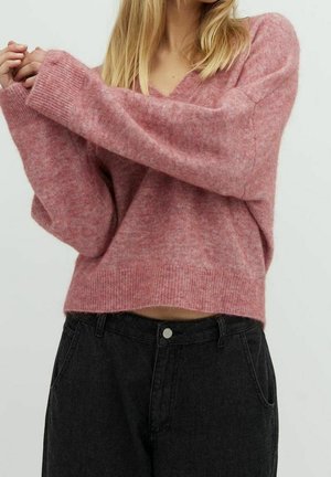 Pull en tricot rose, doux, avec un col en V et des manches amples. Porté avec un pantalon en jean noir taille haute. Le tissu semble duveteux et léger.