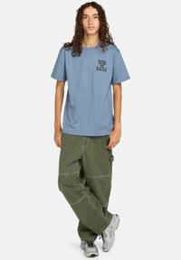 Ung person med langt, krøllet hår iført en blå "Ride the Rails" t-shirt, grønne løse bukser og grå sneakers, stående med korslagte ben.
