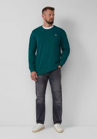 Teal sweatshirt met ribgebreide manchetten en nek, gedragen met versleten donkere jeans en witte sneakers. De sweatshirt heeft een subtiele logo.