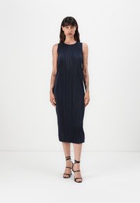 Mekko, tummansinisestä, hihattomasta, laskostetusta midi-mallista, jossa on pyöreä pääntie. Mallilla on mustat, remmillä varustetut korkokengät. Yksinkertainen ja elegantti muotoilu.