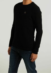 Svart långärmad tröja med ett texturerat mönster, rund halsringning och en liten logotyp på bröstet. Kombineras med mörka jeans.