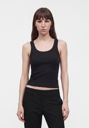 Femme aux longs cheveux bruns portant un haut noir sans manches et un pantalon noir, debout devant un fond clair uni.