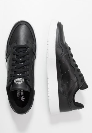 Zapatillas de cuero negro con textura suave, puntera redonda y suela de goma blanca. Presentan cordones negros y detalles perforados en los laterales.