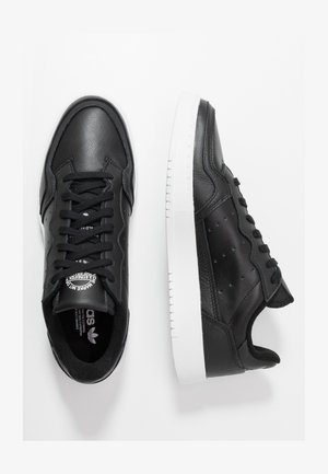 Črne usnjene superge z gladko teksturo, okroglo konico in belo gumijasto podplato. Imajo črne vezalke in perforirane detajle na straneh.