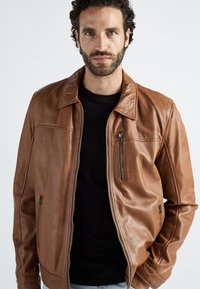 Man met donker krullend haar en baard, gekleed in een bruine leren jas over een zwart shirt, staand tegen een effen witte achtergrond.