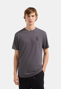 T-shirt grigio a maniche corte in un tessuto morbido con scollatura rotonda e un piccolo logo nero sul lato sinistro, abbinato a pantaloni neri.