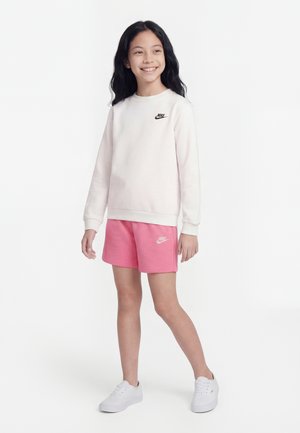 Χαμογελαστή κοπέλα όρθια, φοράει ανοιχτό ροζ φούτερ Nike, ροζ σορτς Nike και λευκά αθλητικά παπούτσια, σε λευκό φόντο.