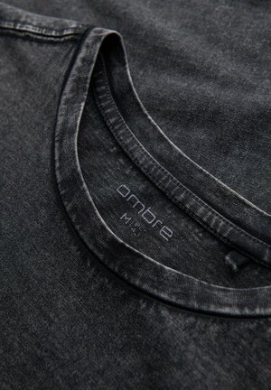 Schwarzes, strukturiertes T-Shirt mit Rundhalsausschnitt, das ein verblasstes Muster aufweist. Das Etikett trägt den Schriftzug "ombre" in dezentem Druck, platziert am inneren Kragen.