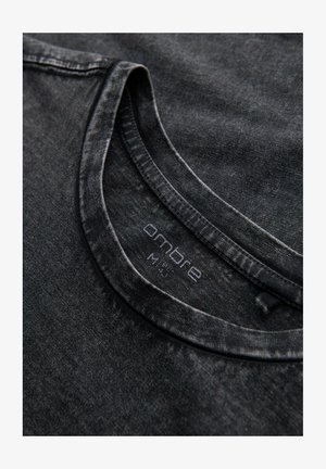 Schwarzes, strukturiertes T-Shirt mit Rundhalsausschnitt, das ein verblasstes Muster aufweist. Das Etikett trägt den Schriftzug "ombre" in dezentem Druck, platziert am inneren Kragen.