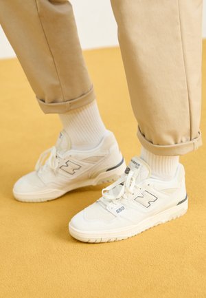 Personne portant un pantalon beige et des baskets New Balance 550 blanches avec des chaussettes blanches, debout sur un sol jaune.