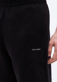 Κοντινό πλάνο μαύρου παντελονιού φόρμας με ελαστική μέση, πλαϊνή τσέπη και μικρό λευκό λογότυπο Calvin Klein κοντά στην τσέπη· το χέρι χαλαρό δίπλα στο παντελόνι.