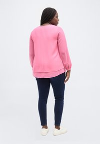 Γυναίκα με πλεξούδες στα μαλλιά, που φοράει ροζ μπλούζα με μακριά μανίκια, σκούρο μπλε τζιν και λευκά αθλητικά παπούτσια, στέκεται πλάτη και κοιτάζει προς τα πίσω σε λευκό φόντο.