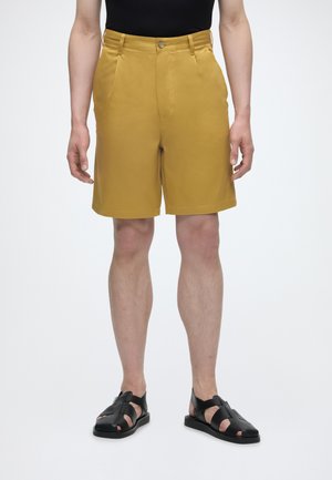 Hombre con pantalones cortos amarillo mostaza, camiseta negra y sandalias de cuero negras, de pie contra un fondo blanco.