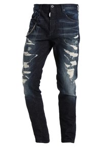 Úzké džíny v tmavě modré barvě s odřenými detaily a bílými skvrnami. Obsahují kapsu na zip a tradiční poutka na opasek.