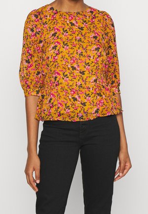 Kvinde iført en sennepsgul bluse med pink blomsterprint og puffede trekvartærmer, kombineret med sorte bukser.