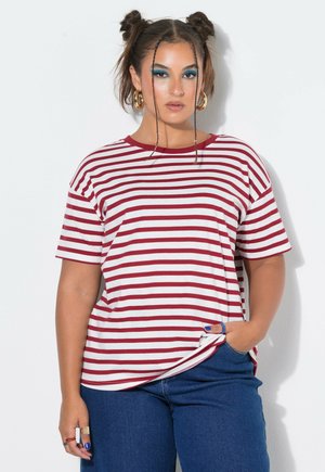 Frau mit zwei geflochtenen Dutts, die ein rot-weiß gestreiftes T-Shirt und blaue Jeans trägt, mit blauem Augen-Make-up und goldenen Creolen.