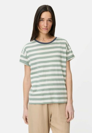 Vrouw met lang, donker haar, gekleed in een groen-wit gestreept shirt met korte mouwen en beige broek, staand tegen een effen achtergrond.