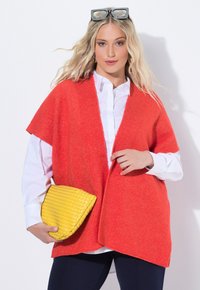 Röd stickad cardigan med breda korta ärmar, bärs över en vit skjorta. Håller en texturerad gul clutchväska, placerad framför en neutral bakgrund.
