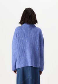 Yli suurikokoinen violetti neulepusero, jossa on korkea kaulus, teksturoitu ribbikuvio, pudotettu olkalinja ja pehmeä, pörröinen materiaali.