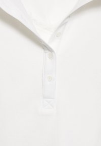 Valkoinen poolopaita, jossa on ribattu pinta, ja siinä on napitettava lista sekä kaulus. Huomiota herättäviä yksityiskohtia ovat pitsikoriste kauluksen alla.