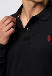 Polo nero con un tessuto texture, caratterizzato da un logo rosso sul petto e un motivo a strisce a contrasto all'interno del colletto.