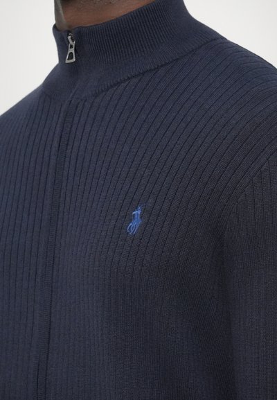 Camisola de malha canelada azul-marinho com gola alta e fecho de meia cremalheira. Apresenta um pequeno logotipo azul no peito e um tecido texturizado.