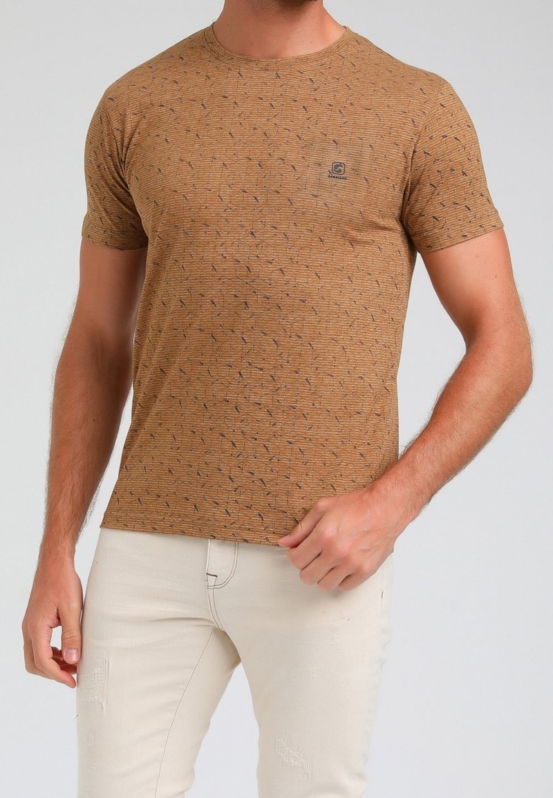 Homme portant un t-shirt à manches courtes marron avec un motif et un pantalon beige clair, debout avec une main près de la taille.