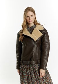Giacca di pelle marrone con colletto in shearling beige. Caratterizzata da una chiusura con zip, tasche laterali e vita con cintura. Indossata sopra un abito floreale a fantasia.