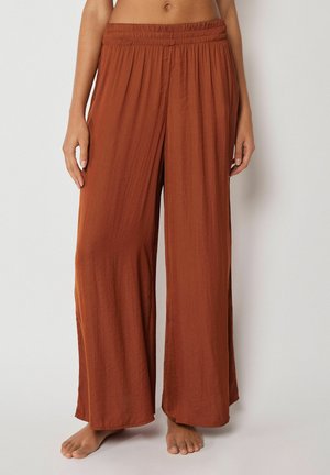 Kvinde iført løstsiddende, vidde bukser i rustfarve med elastik i taljen, stående barfodet mod en ensfarvet baggrund.