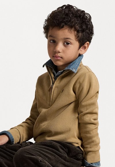 Brun ribbet genser med glidelås krage, lagd over en denimblå skjorte. Barnet sitter og har på seg mørke kordfløyelsbukser.