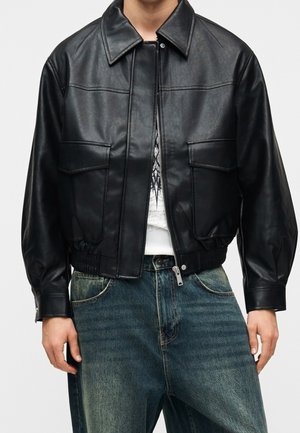 Personne portant une veste en cuir noire avec poches avant à rabat, taille élastique et jean bleu, montrée du cou aux hanches.