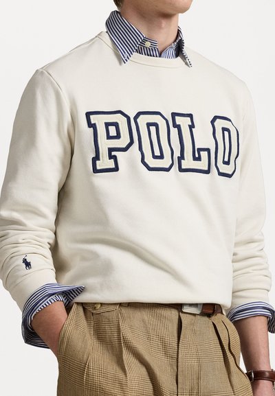 Sweatshirt crem cu broderie albastră "POLO", purtat peste o cămașă cu dungi alb-albastre, asortat cu pantaloni bej cu un model în carouri.