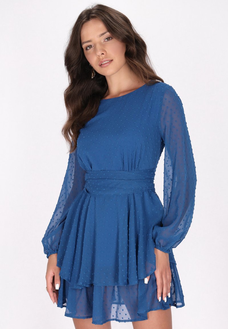 Rochie albastră dintr-un material ușor, texturat, cu un model punctat. Dispune de mâneci lungi dintr-un material transparent și de un design cu fusta în straturi, evazată.