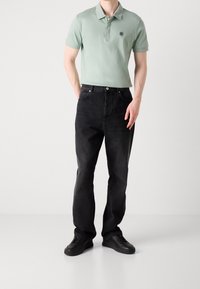 Camiseta polo de manga corta en verde claro con cuello y logo, combinada con pantalones vaqueros negros de pierna recta y zapatillas negras. Textura de tejido suave.